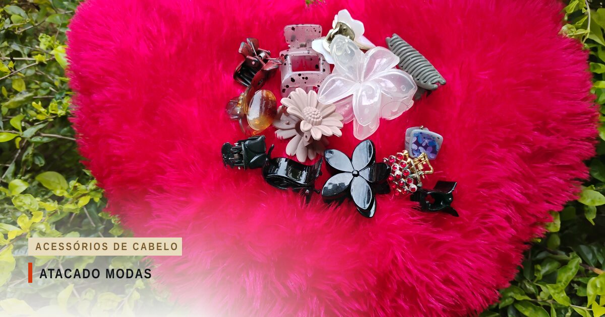 Presilhas de cabelo variadas, entre elas garras em forma de flor, nas cores preta, bege, branca e translúcida, sobre uma almofada felpuda cor-de-rosa em forma de coração, cercada de folhagem