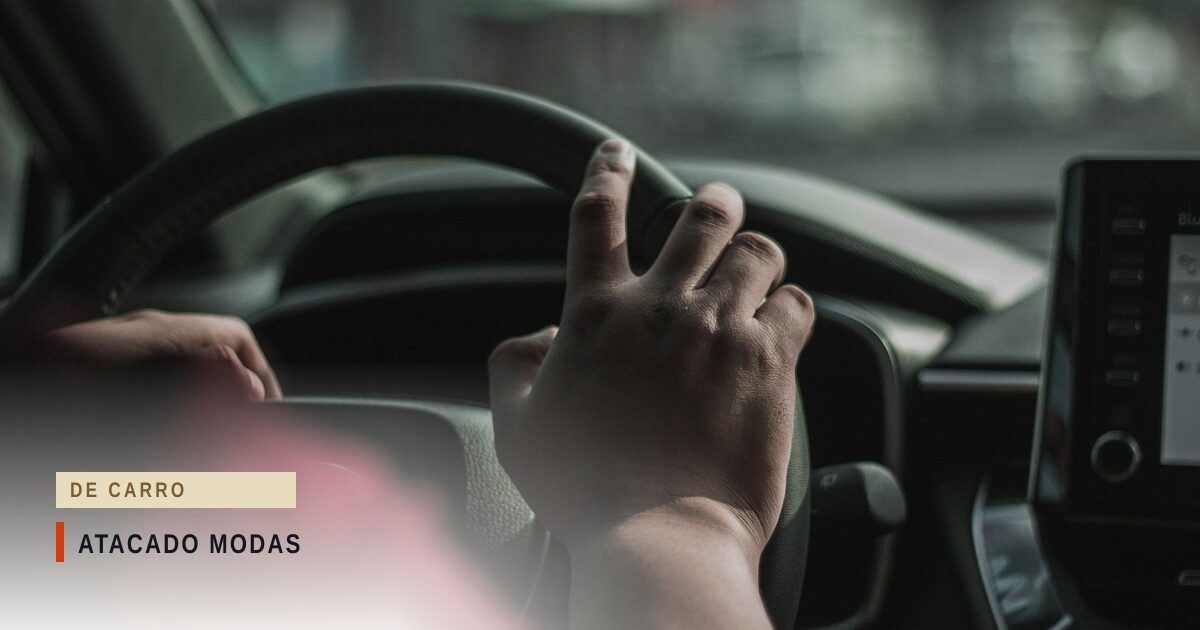 Mãos de um motorista no volante de um carro, com o painel à direita e a rua desfocada pelo para-brisa