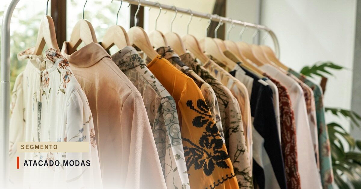 Arara branca com blusas femininas em cabides de madeira — floral, rosa liso, mostarda com bordado preto e azul-marinho — diante de uma janela com folhagem ao fundo