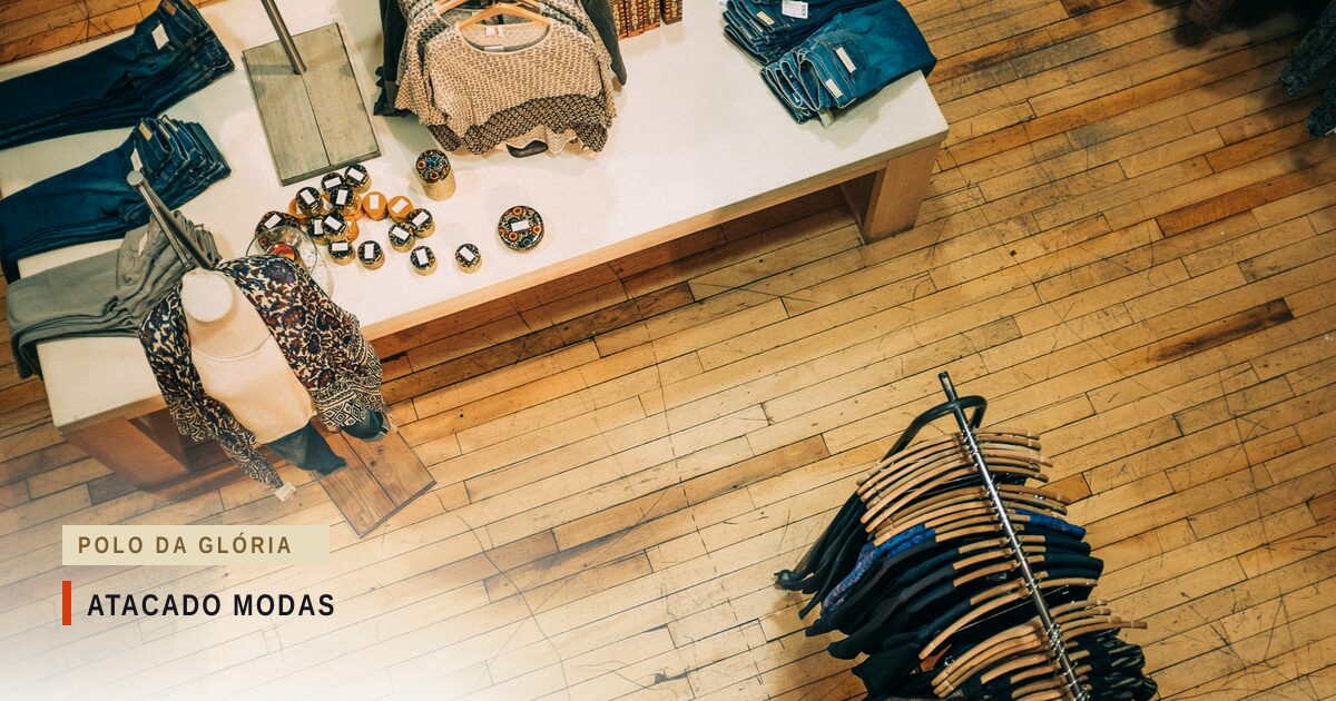 Loja de roupa vista de cima: mesa branca com calças jeans dobradas, blusas e acessórios, um manequim de busto com lenço estampado e uma arara de cabides com peças escuras sobre o piso de madeira