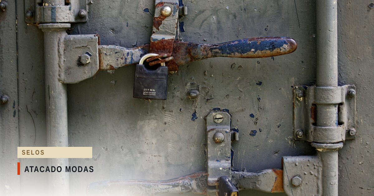 Close das portas metálicas de uma carga fechada, com barras de travamento horizontais enferrujadas, tinta cinza descascada e um cadeado de metal pendurado na trava superior