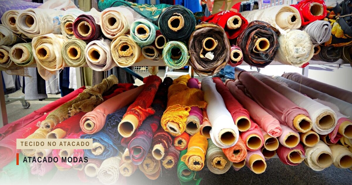 Rolos de tecido de várias cores, empilhados numa prateleira de loja e deitados sobre uma bancada, com o tubo de papelão à mostra nas pontas