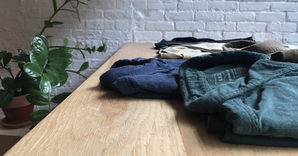 Peças de roupa dobradas sobre uma mesa de madeira clara, com parede de tijolo pintada de branco e um vaso de planta ao lado