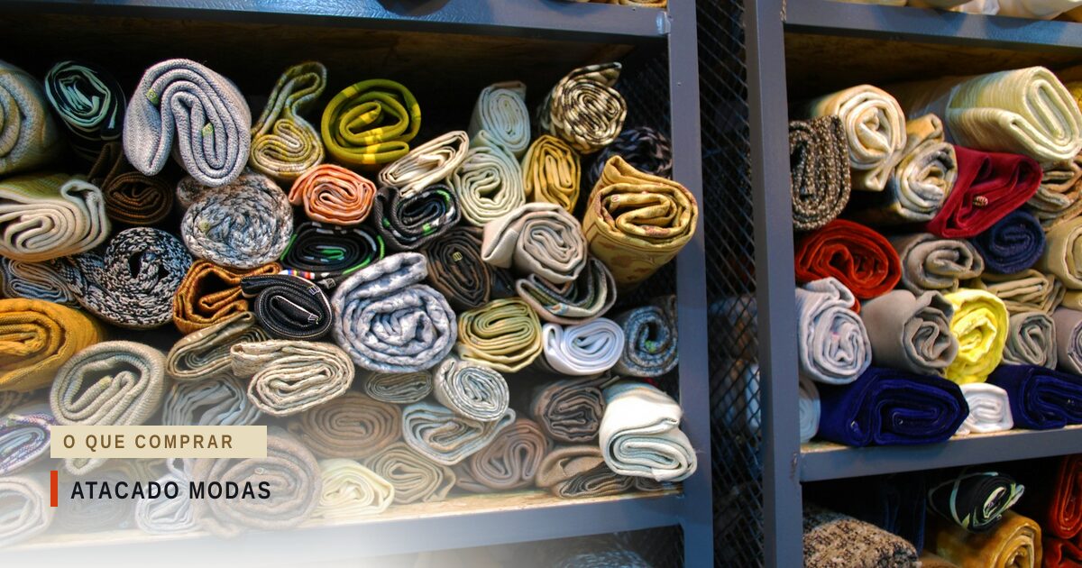 Estantes de metal cinza abarrotadas de peças de malha e tecido enroladas e dobradas, em dezenas de cores e padronagens, empilhadas até o topo de cada prateleira